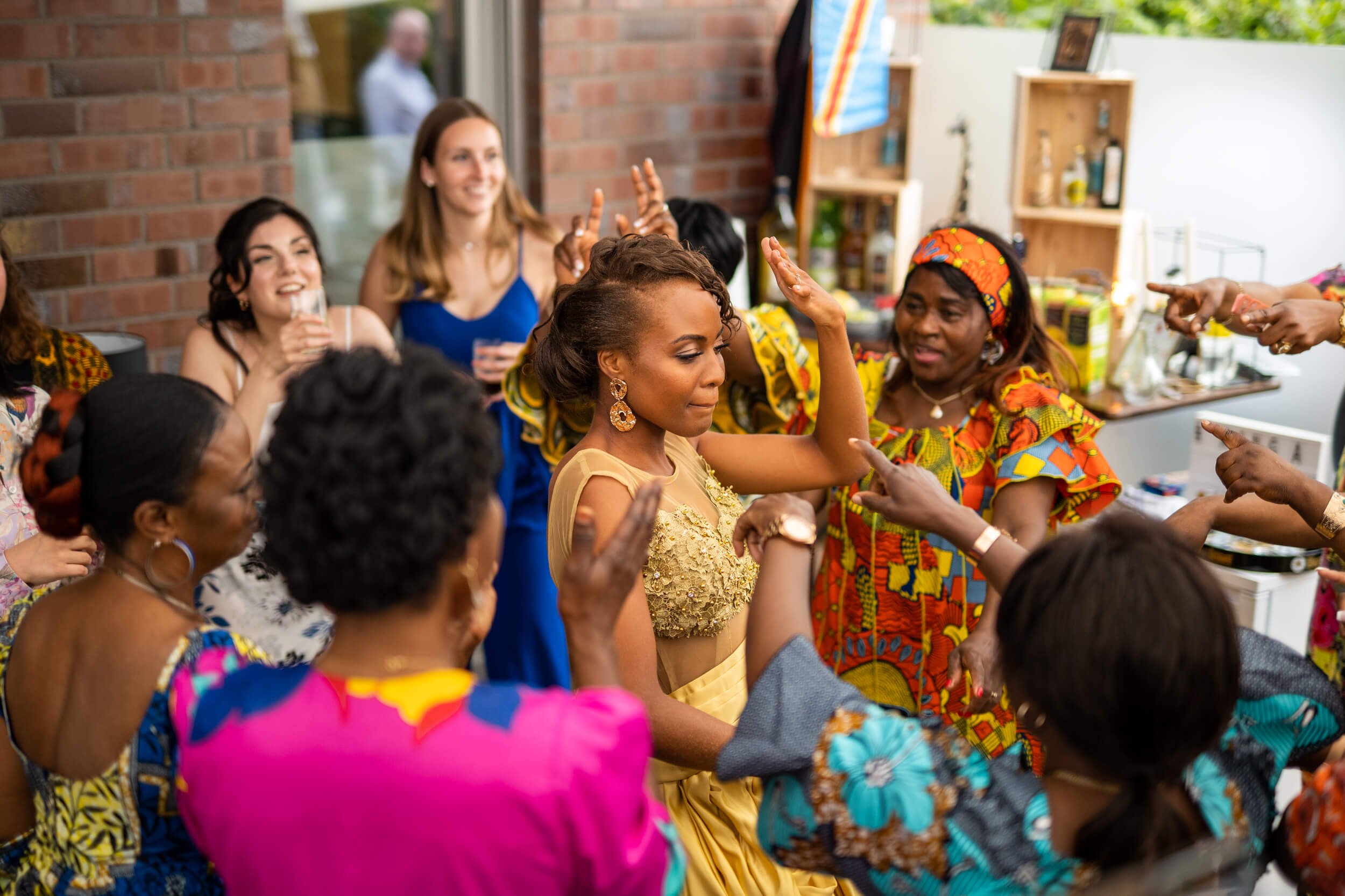 Feiernde Frauen in bunten Kleidern umringt eine Dame in einem goldenen Kleid. Fröhliche Stimmung.