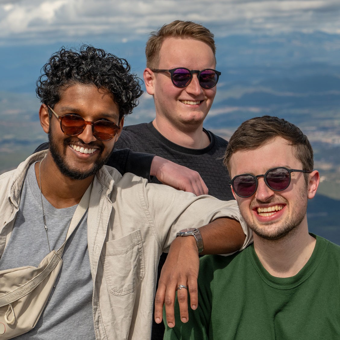 Drei junge Männer mit Sonnenbrillen lächeln glücklich vor einer schönen Landschaft.