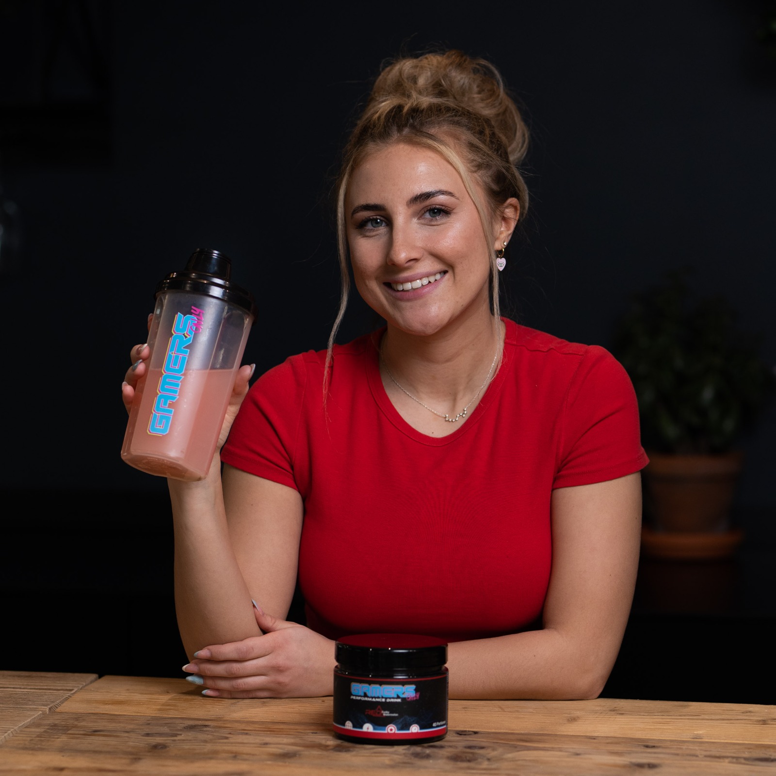 Eine Frau in rotem Shirt hält einen Shaker mit pinkem Getränk und steht neben einem Produkten.