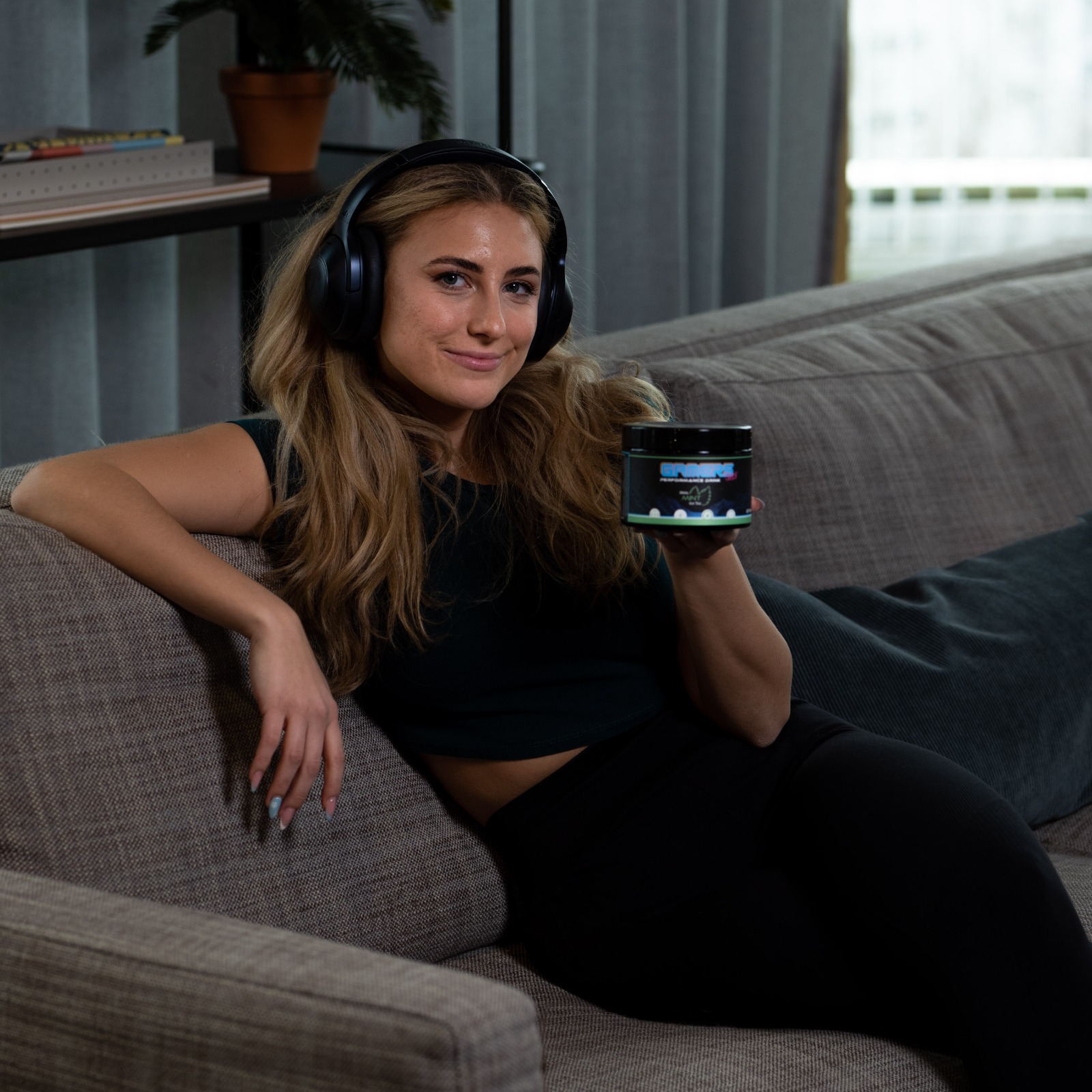 Junge Frau mit Kopfhörern hält ein Produkt in der Hand und sitzt gemütlich auf einem Sofa.