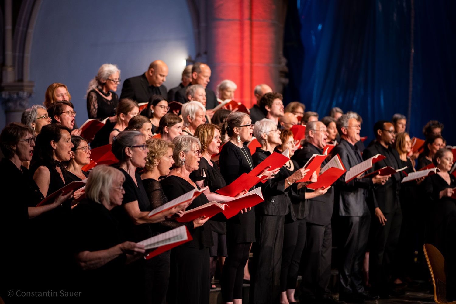 Ein gemischter Chor singt leidenschaftlich mit roten Notenblättern in der Hand.