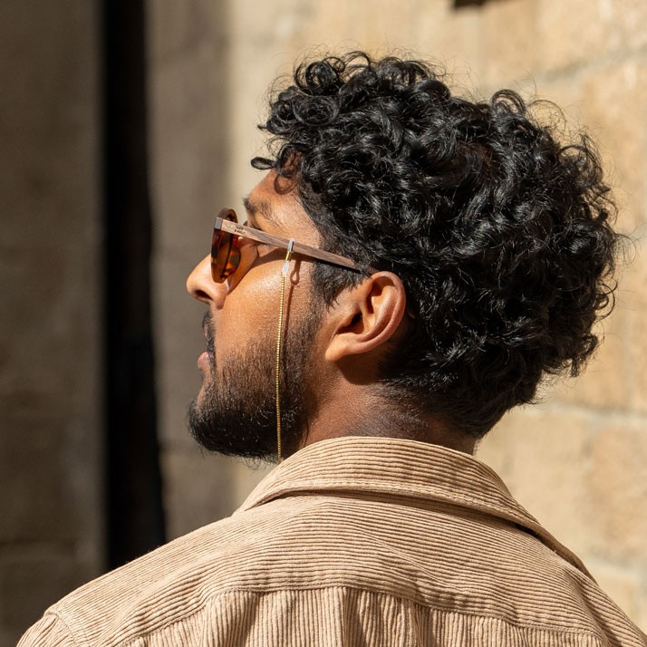 Junger Mann mit lockigem Haar und Sonnenbrille, Blick zur Seite, vor steinerner Wand.