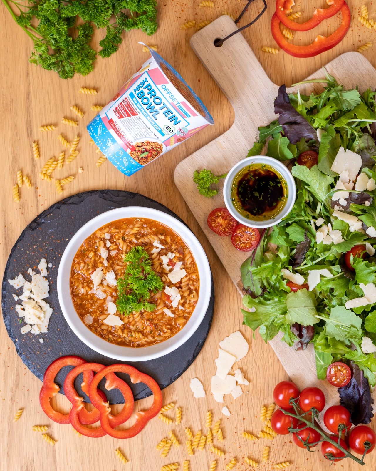 Bunte Mahlzeit mit Pasta, Salat, Tomaten und einer Protein-Bowl auf Holzunterlage.
