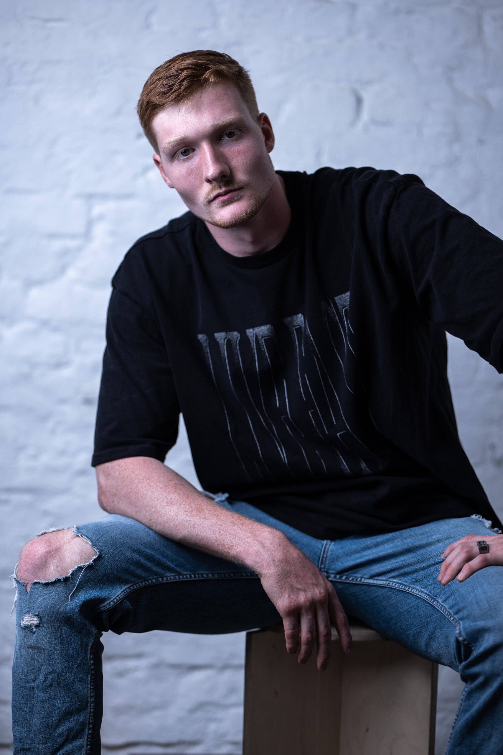Junger Mann mit rotem Haar sitzt lässig in Jeans und schwarzem T-Shirt vor einer hellen Wand.