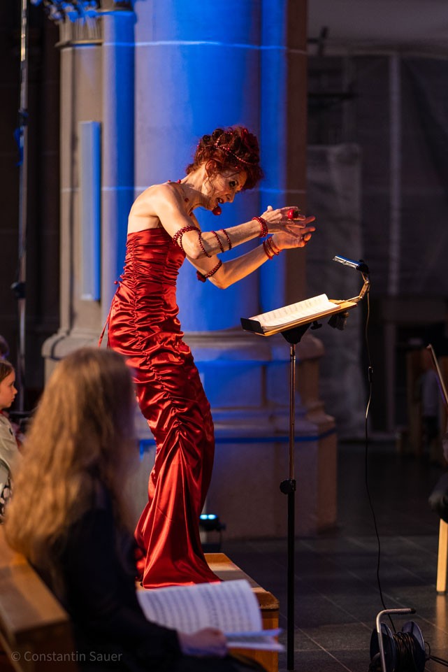 Eine Künstlerin in rotem Kleid performt leidenschaftlich vor Publikum.