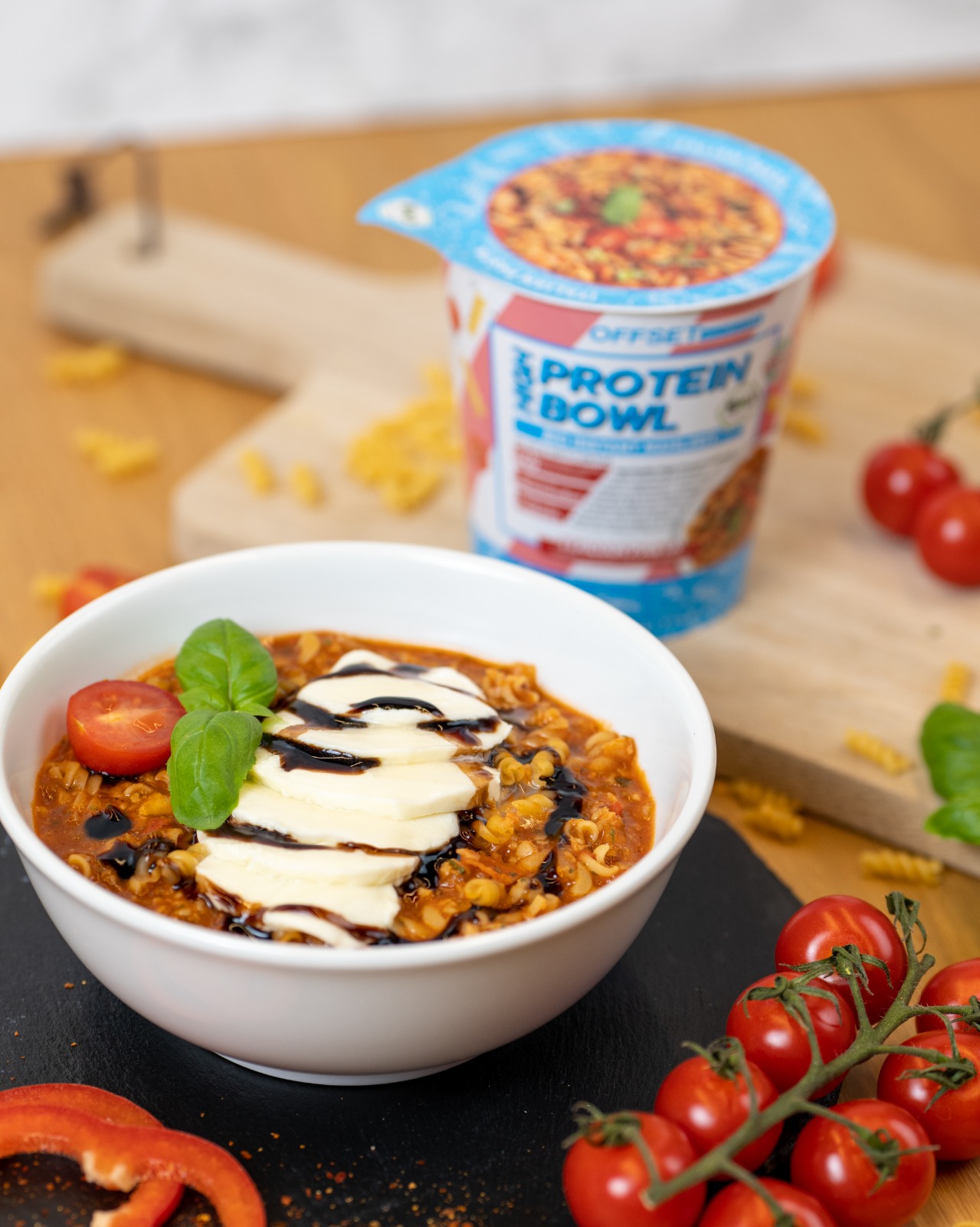 Schale mit Pasta in Sauce, garniert mit Käse, Basilikum und Tomaten, daneben eine Verpackung.
