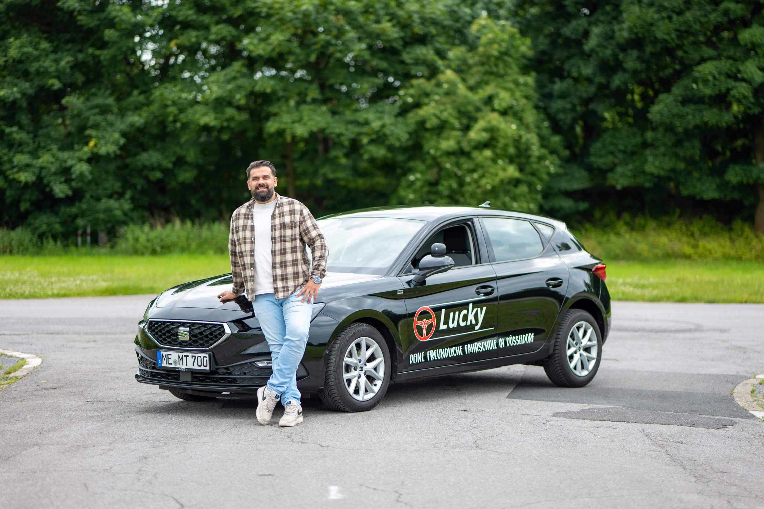 Ein Mann lehnt an einem schwarzen Auto mit der Aufschrift "Lucky" in einer grünen Umgebung.