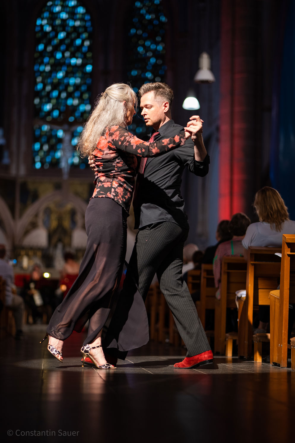 Tanzendes Paar in eleganter Kleidung vor bunten Kirchenfenstern.