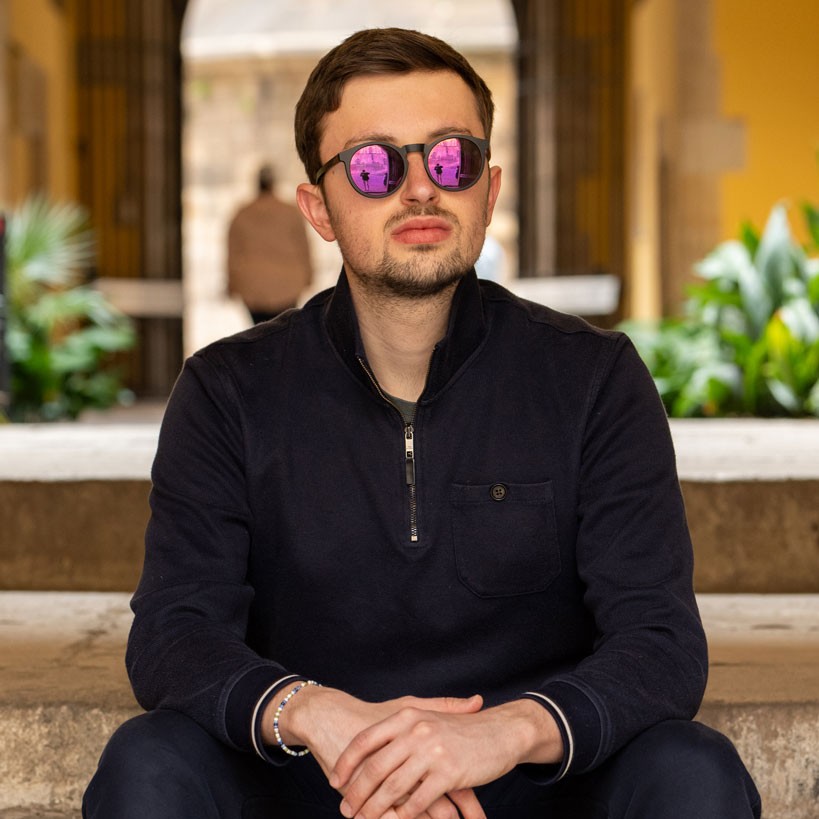 Junger Mann mit Sonnenbrille sitzt selbstbewusst in einer architektonischen Umgebung.
