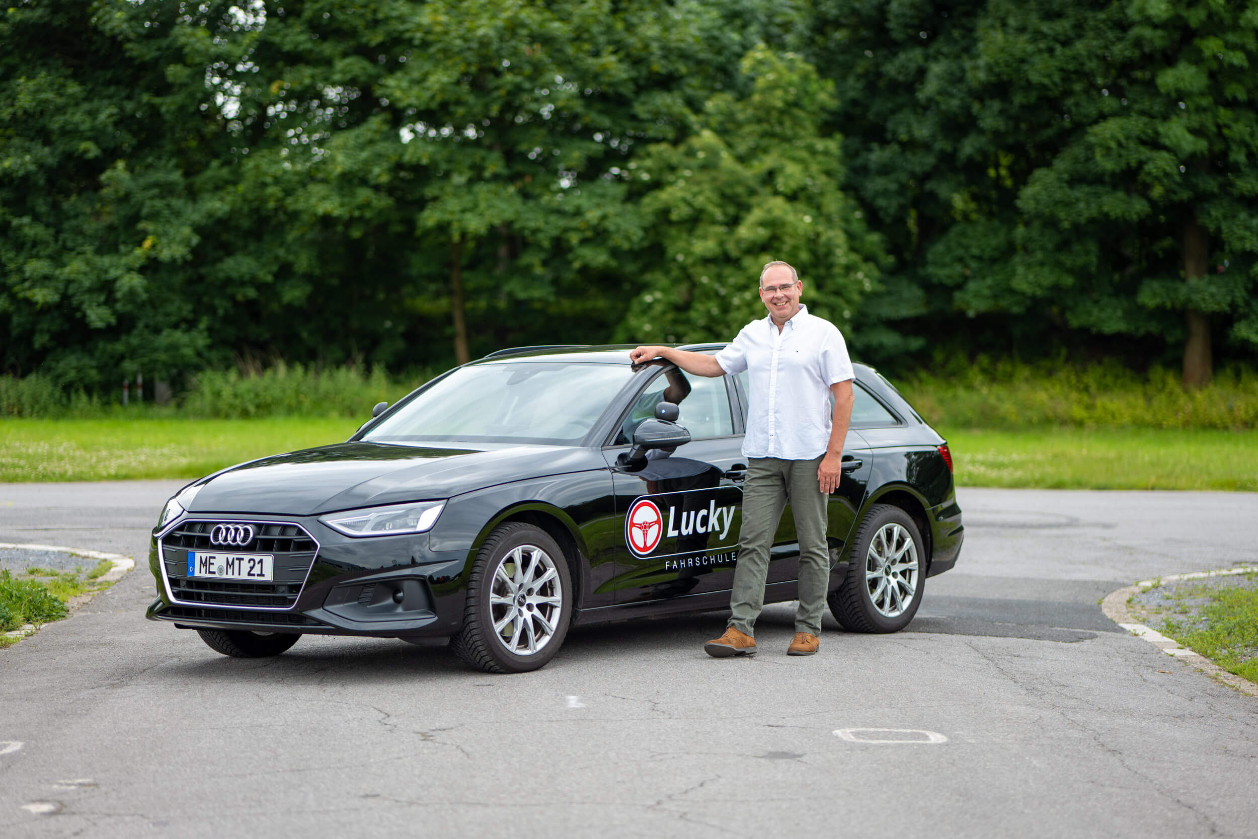 Mann steht neben einem schwarzen Audi mit Lucky-Fahrschule-Logo in einem grünen Park.